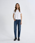 A woman stands against a light gray studio background wearing a plain white short-sleeve T-shirt, blue straight-leg jeans, and black ankle boots.