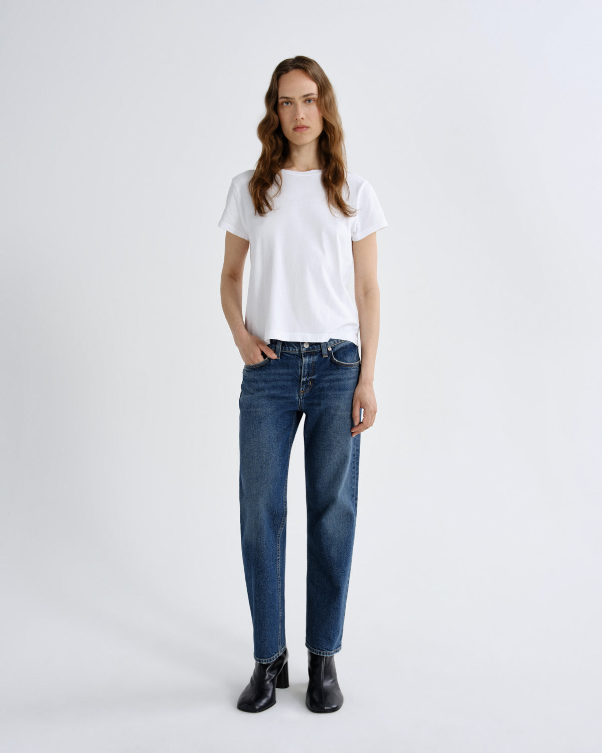 A woman stands against a light gray studio background wearing a plain white short-sleeve T-shirt, blue straight-leg jeans, and black ankle boots.
