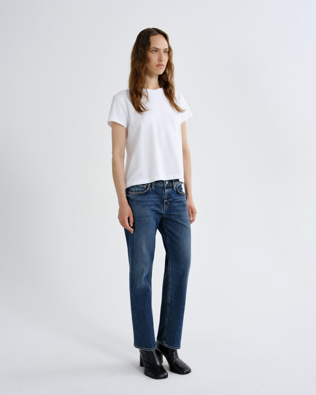 A woman modeling a white short-sleeve crewneck t-shirt and dark blue straight-leg jeans with black ankle boots, standing against a light gray studio background.