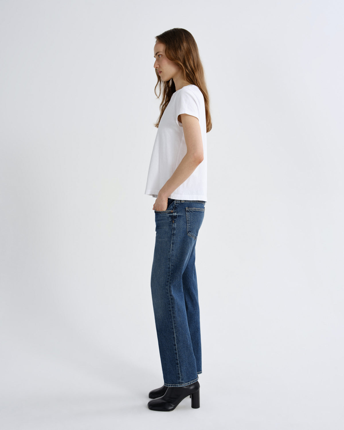 A woman standing in profile wearing a white short-sleeve t-shirt, blue straight-leg jeans, and black heeled boots against a light gray studio background.
