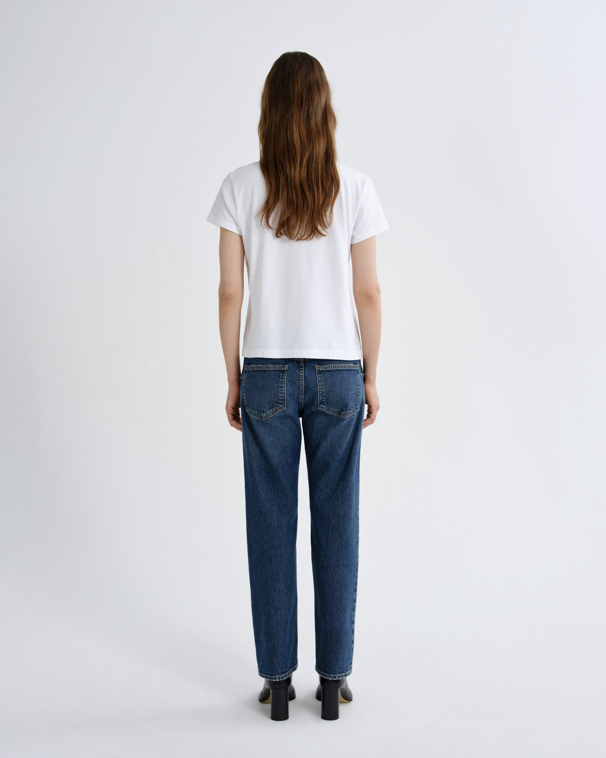 A woman viewed from behind wearing a white short-sleeve t-shirt, blue straight-leg jeans, and black block-heeled shoes against a light gray studio background.