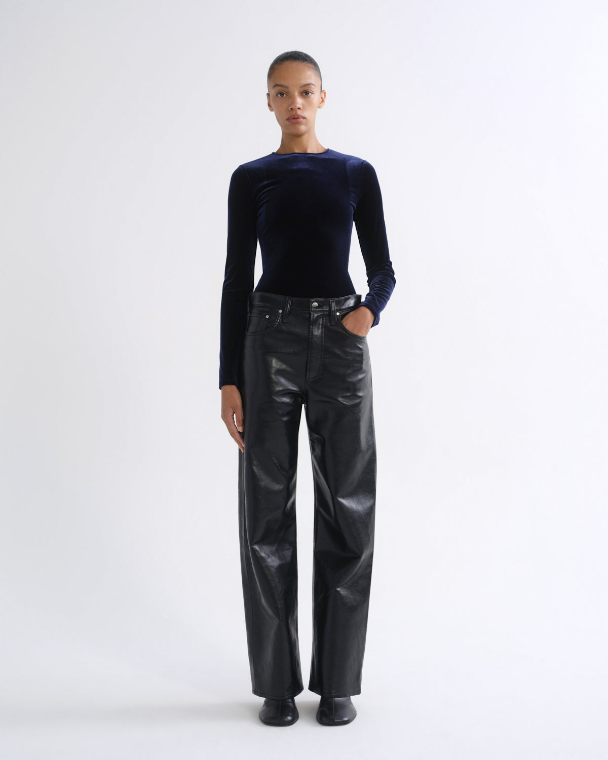 A model stands against a plain light gray studio background wearing a dark navy long-sleeve velvet top and shiny black wide-leg pants.