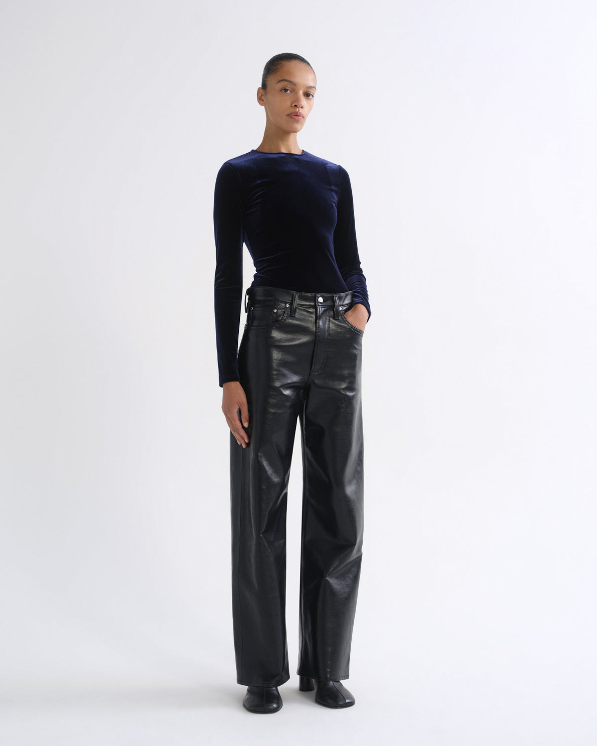 A model stands against a plain white studio background wearing a dark navy long-sleeve velvet top and high-waisted black wide-leg faux-leather trousers.