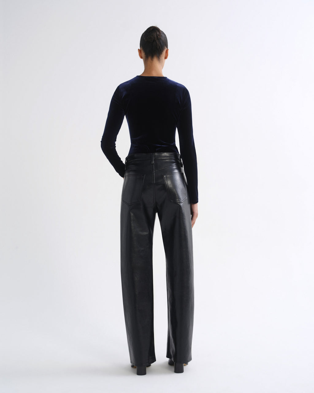 A model shown from the back wearing a black velvet long-sleeve top and black wide-leg leather trousers against a plain white studio background.