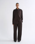 A model stands against a light gray studio background wearing a dark brown long-sleeve button-up shirt and matching dark brown wide-leg trousers.