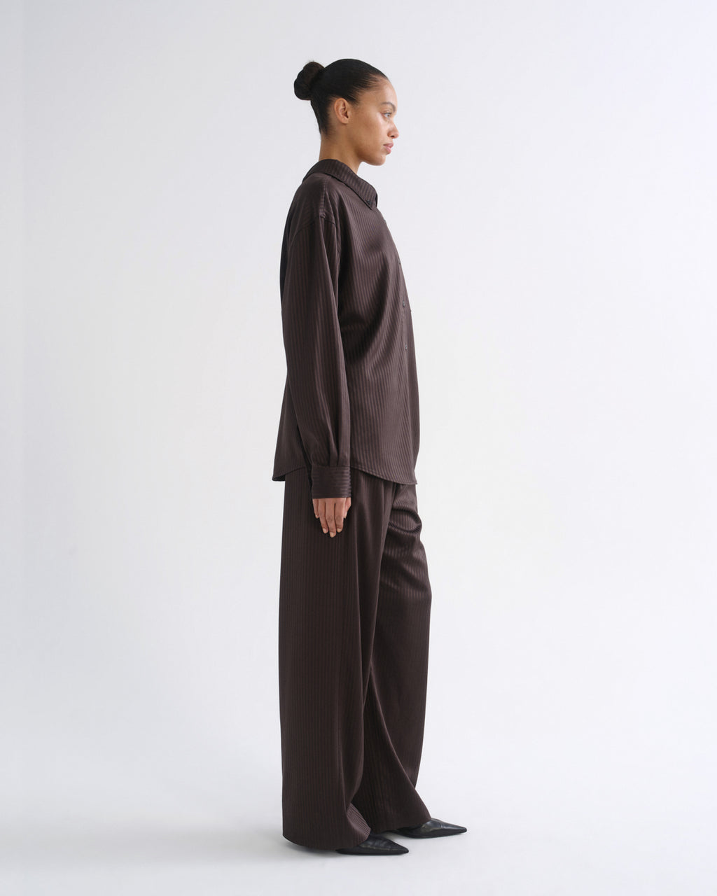 A model in side profile wearing a dark brown long-sleeve button-up shirt and matching wide-leg trousers against a plain white studio background.