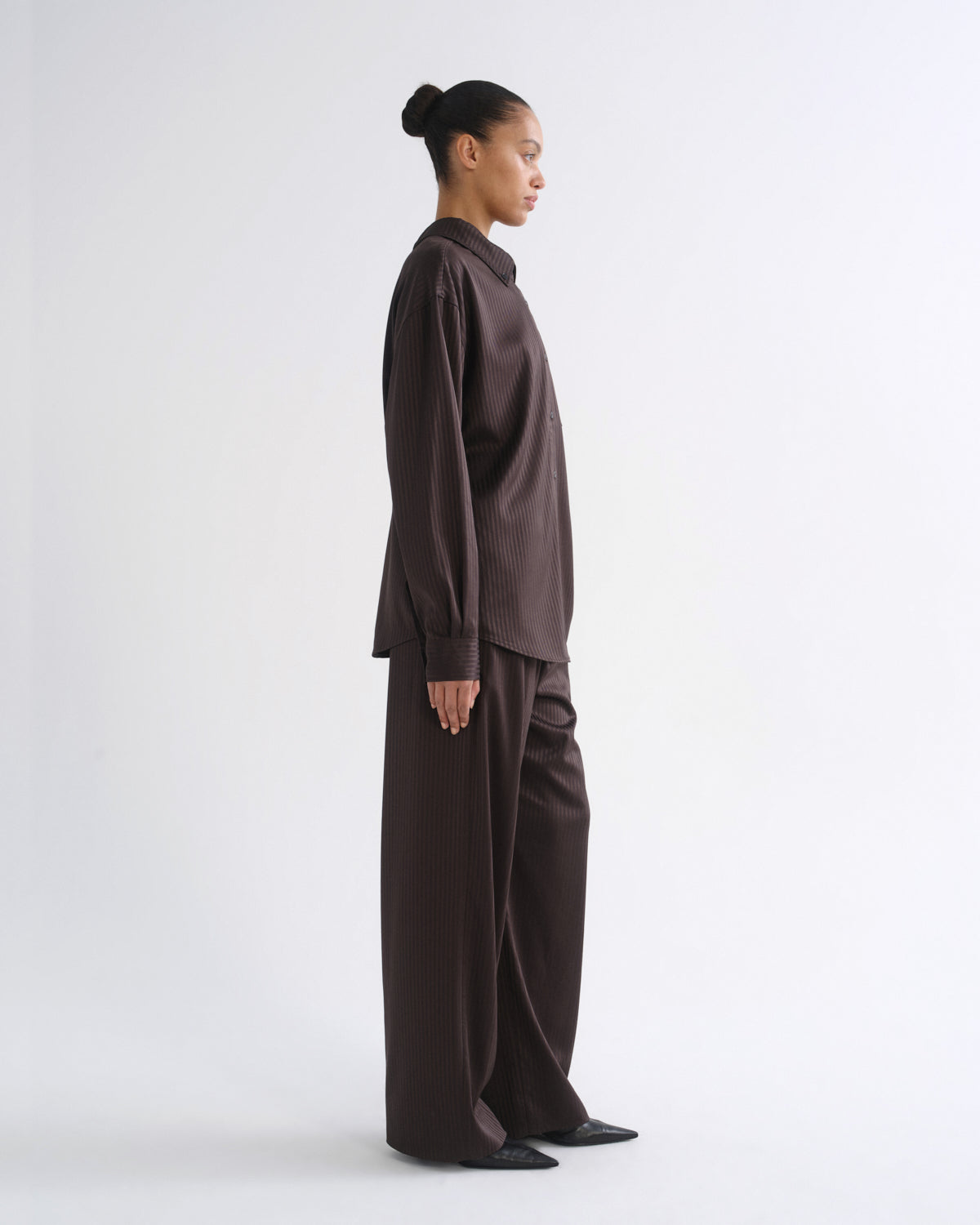 A model in side profile wearing a dark brown long-sleeve button-up shirt and matching wide-leg trousers against a plain white studio background.