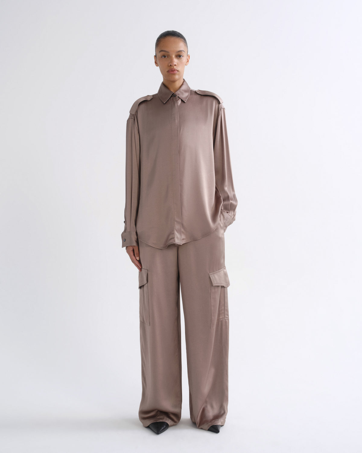 A model stands against a plain white studio background wearing a matching two-piece outfit consisting of a long-sleeve collared button-up shirt and wide-leg cargo pants in a muted taupe-brown color.