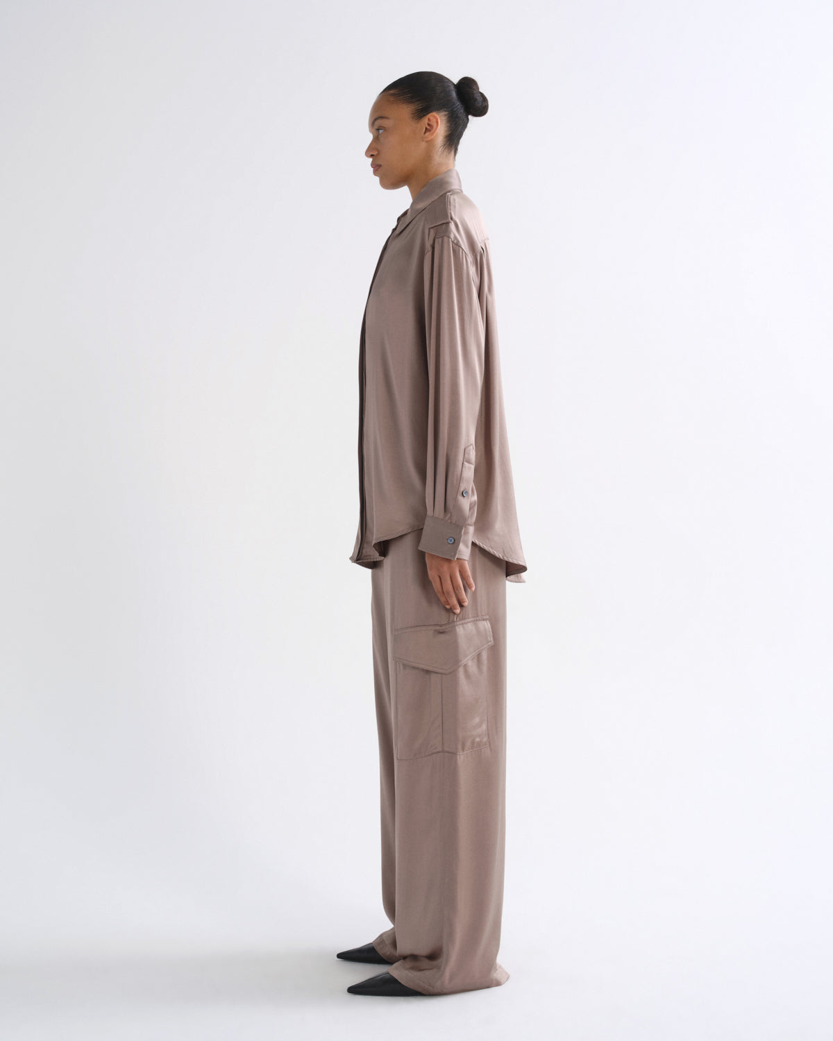 A model in side profile wearing a matching taupe-brown long-sleeve button-up shirt and wide-leg cargo pants set, photographed against a seamless white studio background.
