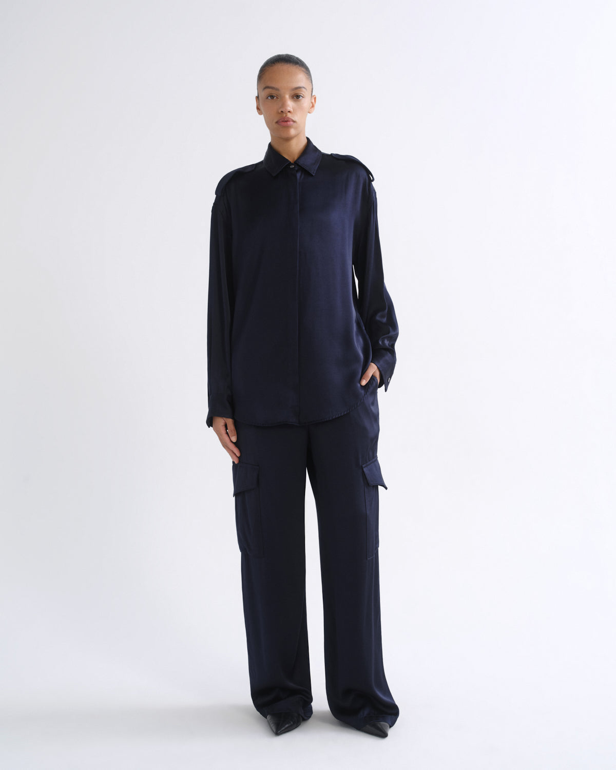 A model stands against a plain white studio background wearing a dark navy blue long-sleeve collared shirt and matching wide-leg cargo pants.