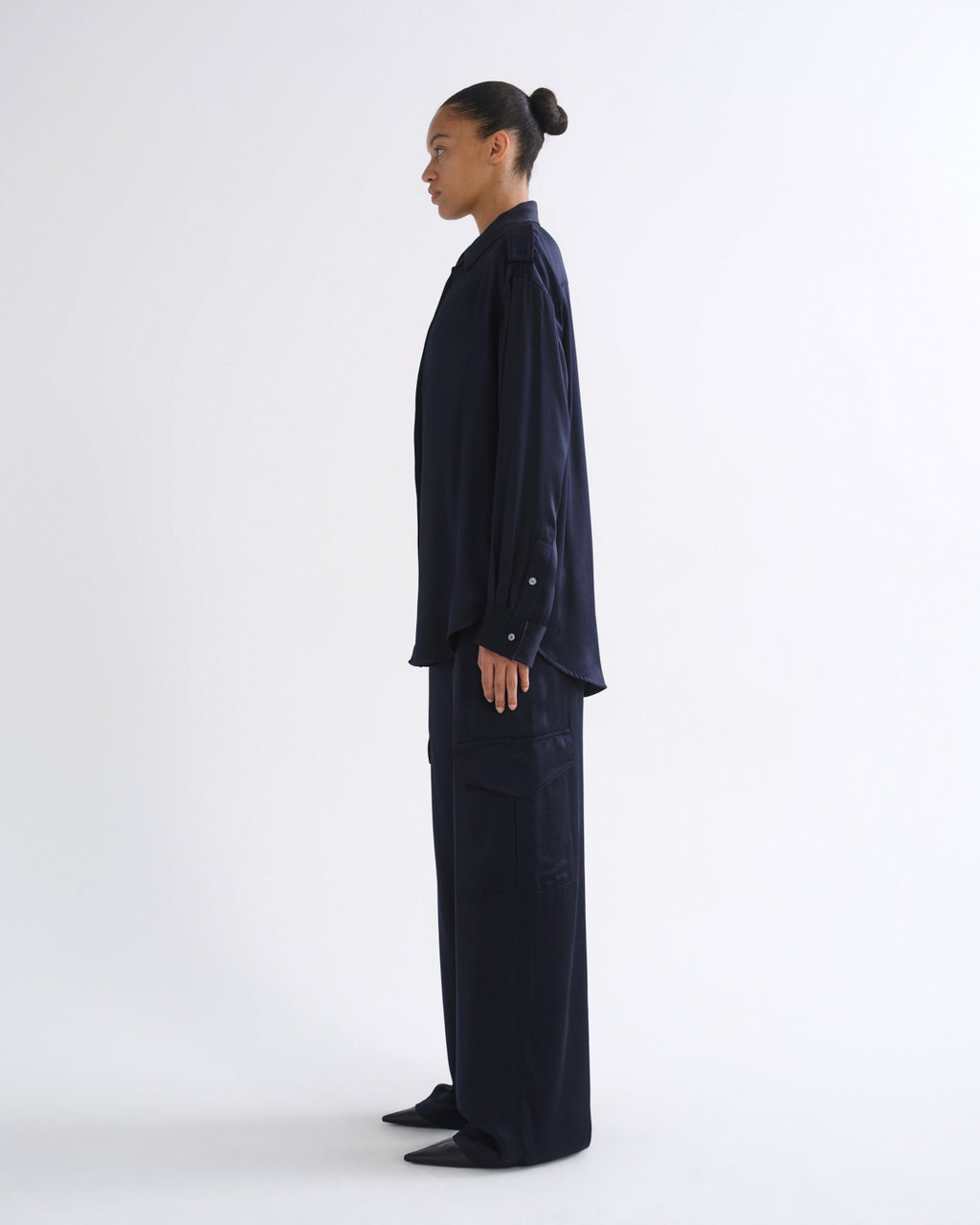 A model in side profile wearing a dark navy blue long-sleeve button-up shirt and matching wide-leg trousers, standing against a plain white studio background.