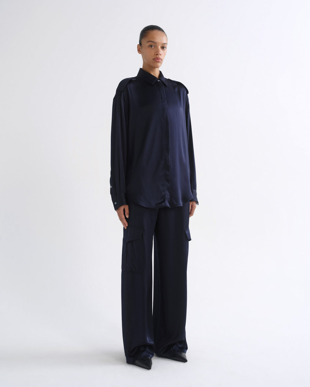 A model stands against a plain white studio background wearing a dark navy blue satin long-sleeve button-up shirt and matching wide-leg trousers.