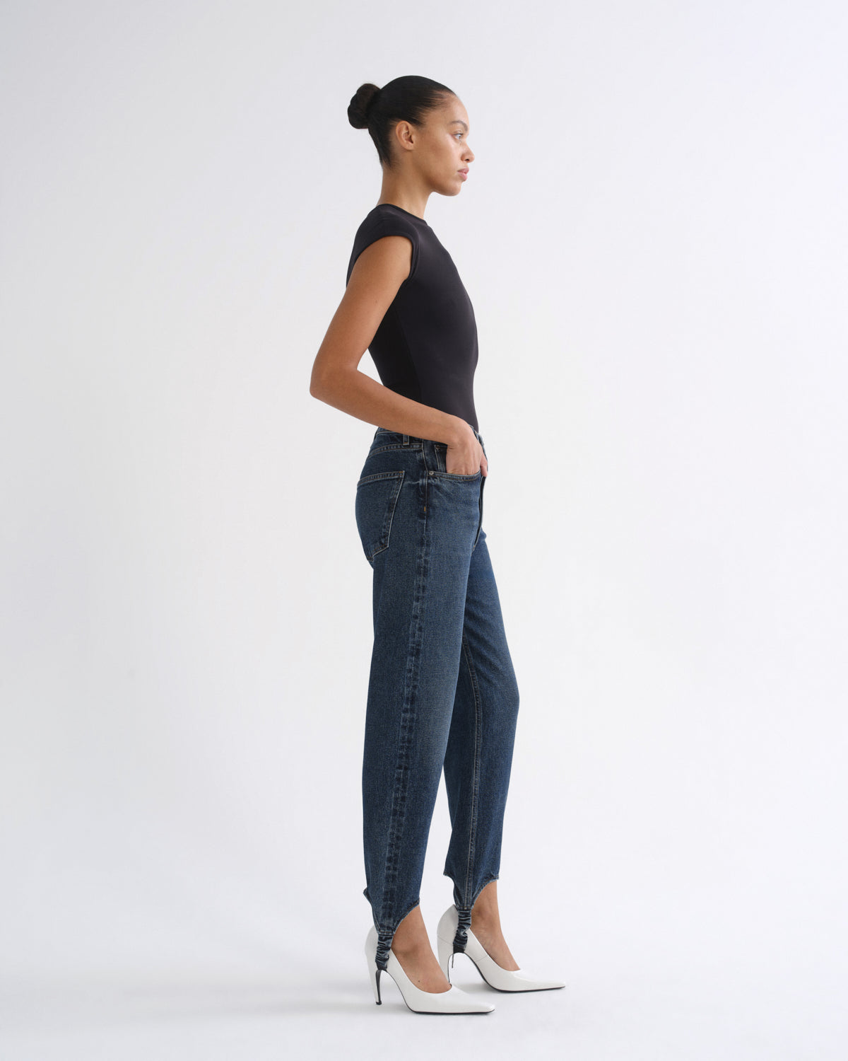 Woman wearing a black sleeveless top with dark indigo straight leg jeans viewed from the side on a grey background.