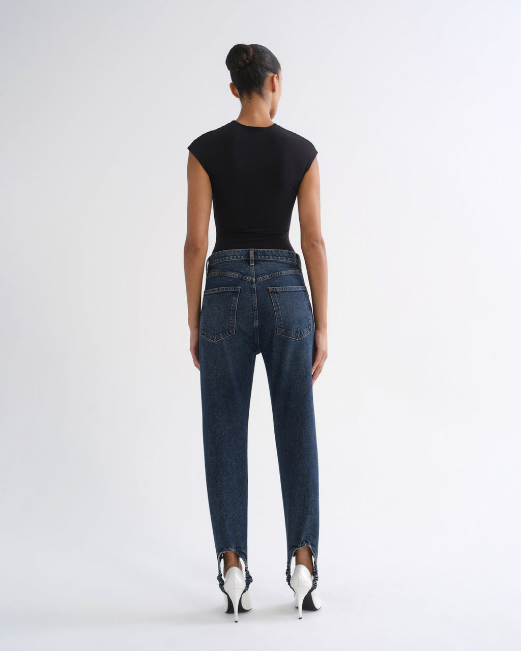 A model shown from the back wearing a black short-sleeve top, dark blue high-waisted straight-leg jeans, and white pointed-toe heels, set against a plain light gray studio background.