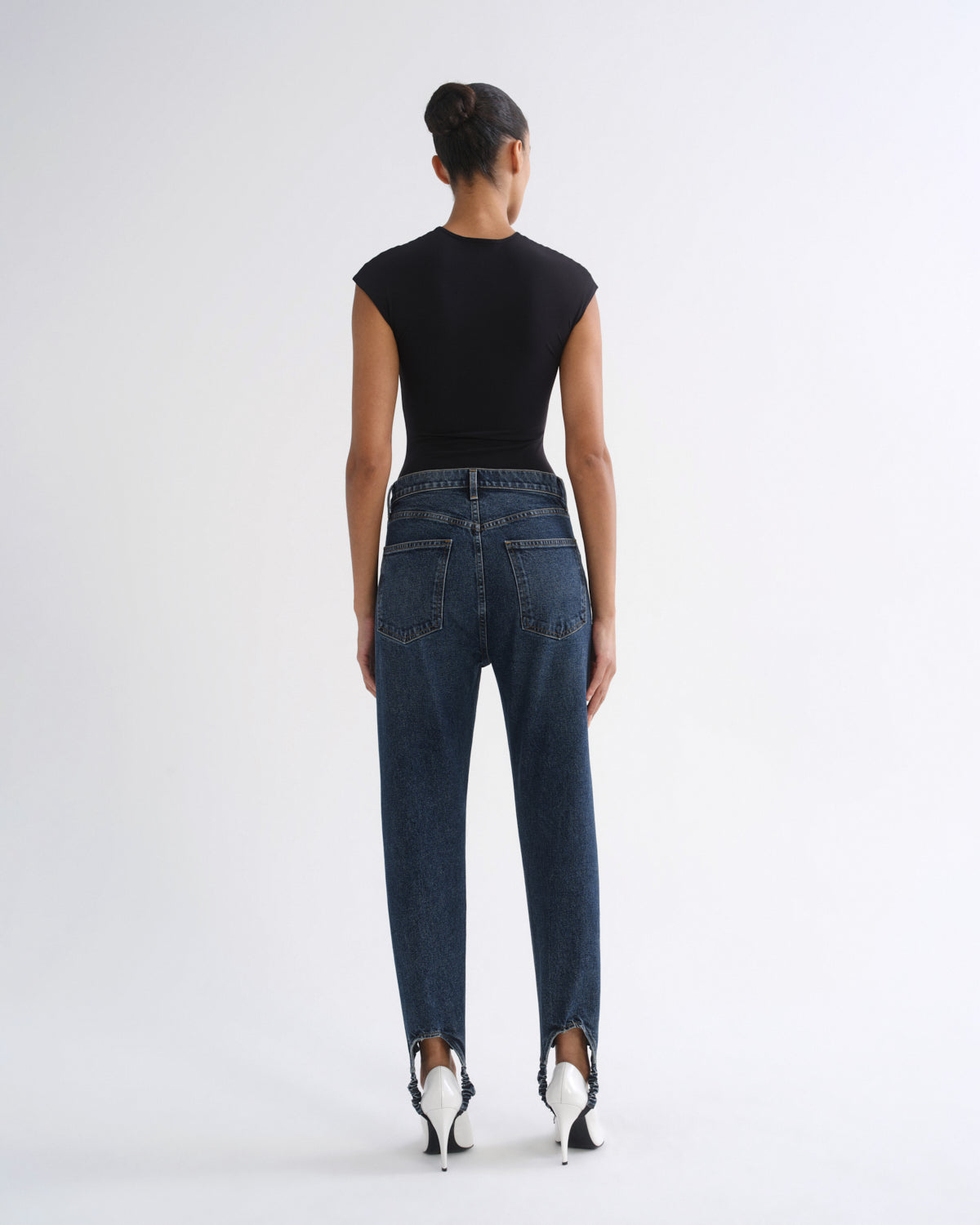 A model shown from the back wearing a black short-sleeve top, dark blue high-waisted straight-leg jeans, and white pointed-toe heels, set against a plain light gray studio background.
