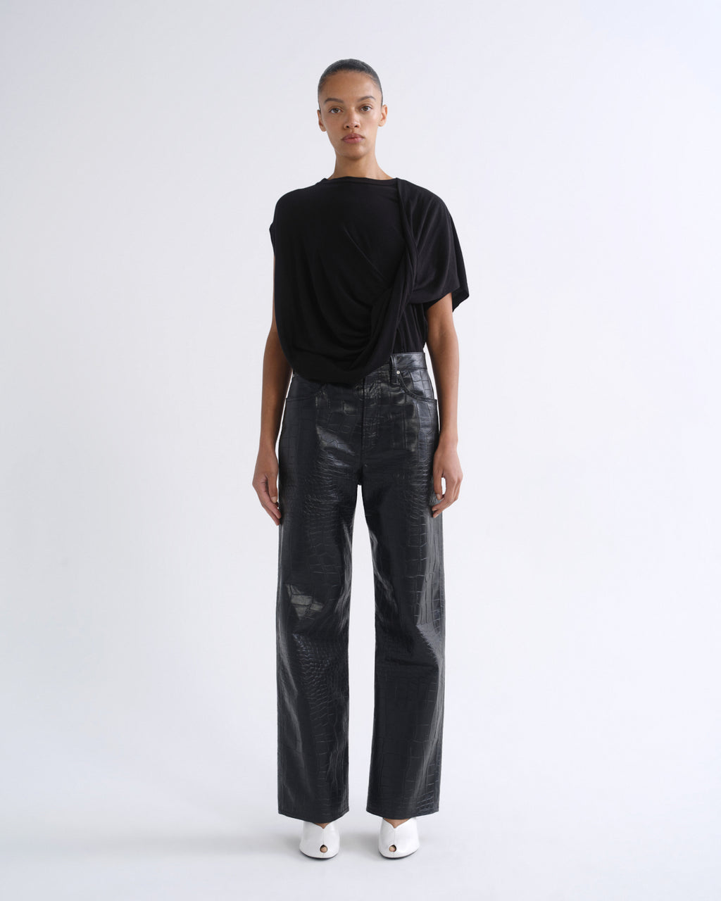A model stands against a plain white studio background wearing a black draped short-sleeve top, black wide-leg pants with a glossy textured finish, and white pointed shoes.