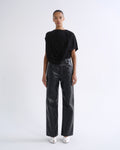 A model stands against a plain white studio background wearing a black draped short-sleeve top, black wide-leg pants with a glossy textured finish, and white pointed shoes.