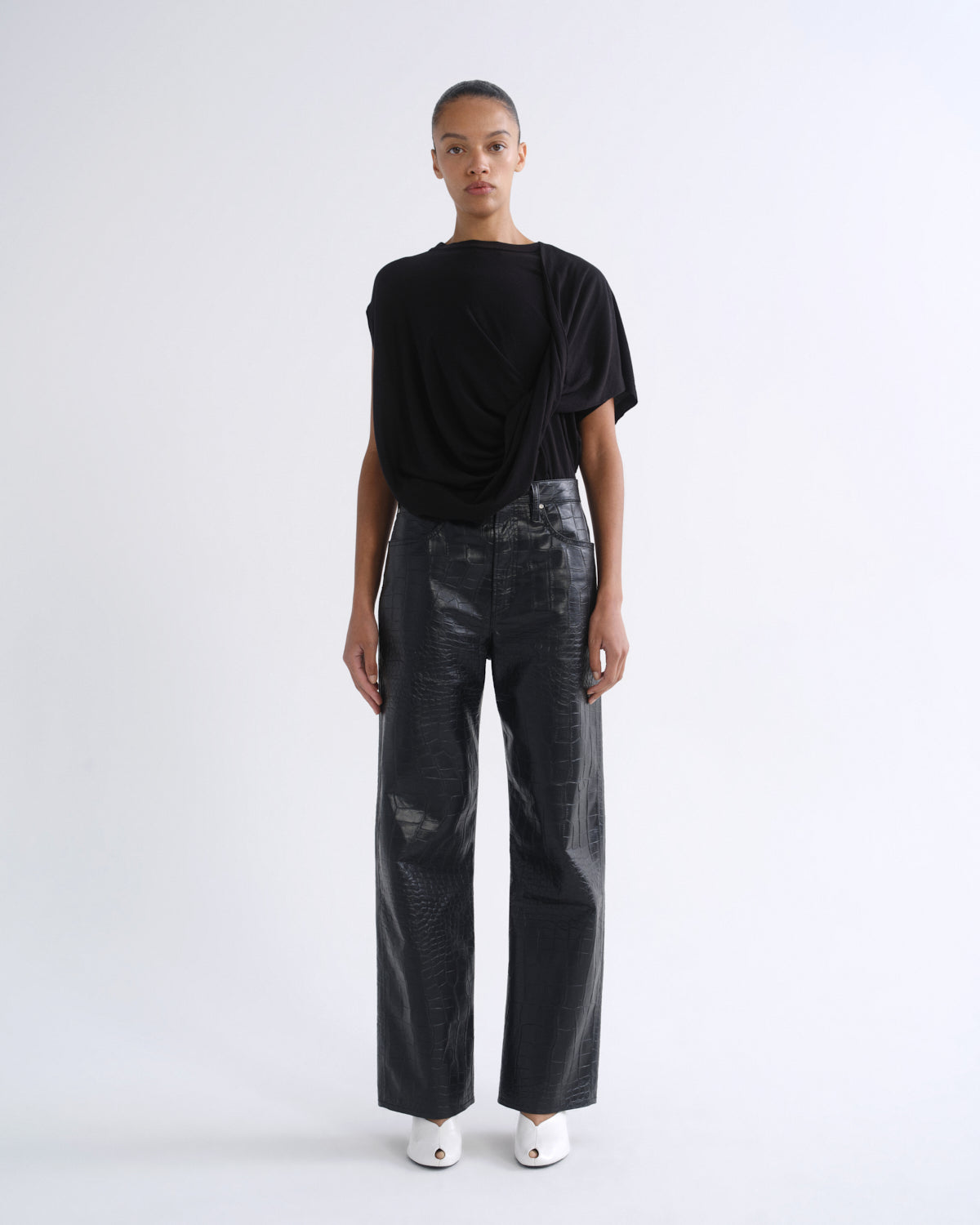 A model stands against a plain white studio background wearing a black draped short-sleeve top, black wide-leg pants with a glossy textured finish, and white pointed shoes.