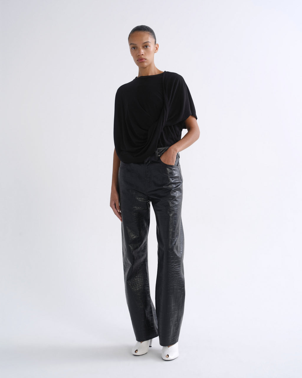 A model stands against a plain white studio background wearing a black draped short-sleeve top, black wide-leg pants with a glossy textured finish, and white pointed shoes.