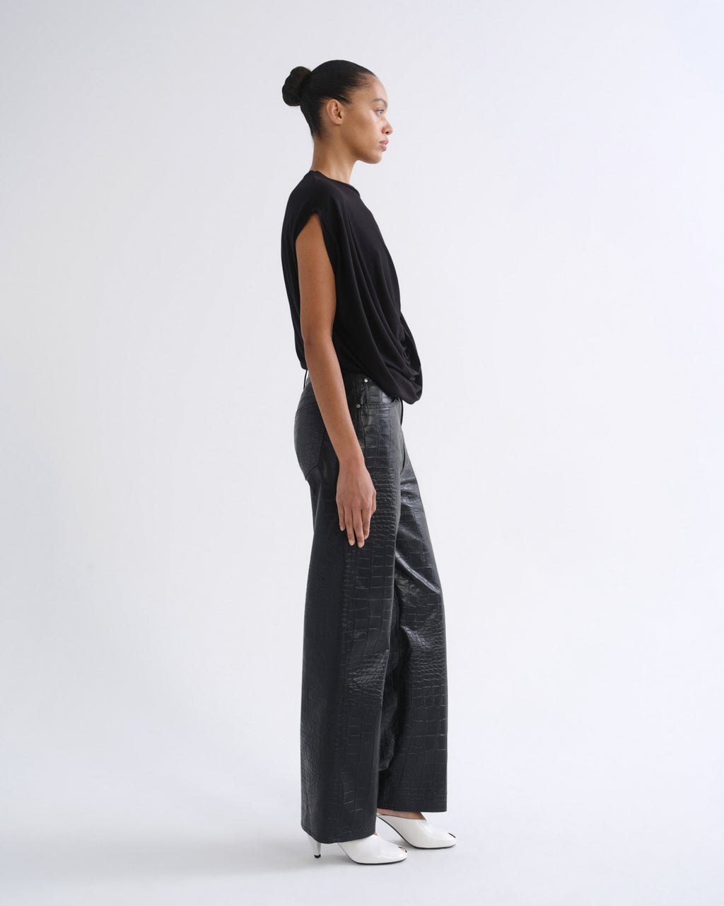 A model is shown in profile wearing a black draped sleeveless top, textured dark gray wide-leg pants, and white pointed-toe heels against a plain white studio background.