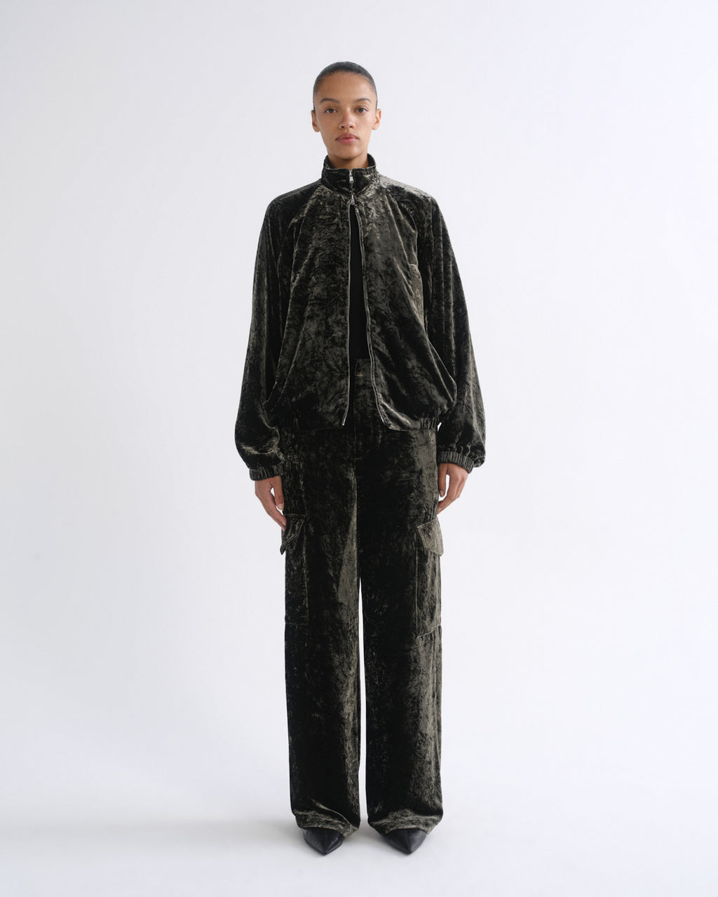 A model stands against a light gray studio background wearing a matching black velvet tracksuit with a high collar zip jacket and wide-leg cargo pants.