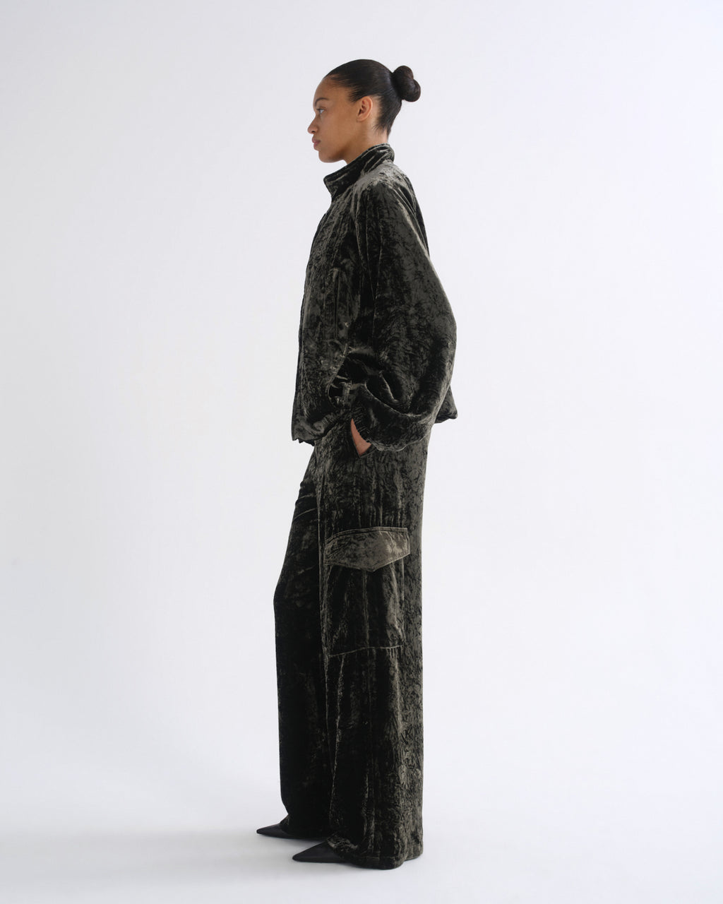 A model in profile wearing a dark charcoal gray velvet two-piece lounge set, featuring a cropped long-sleeve top and matching wide-leg trousers, against a plain white studio background.