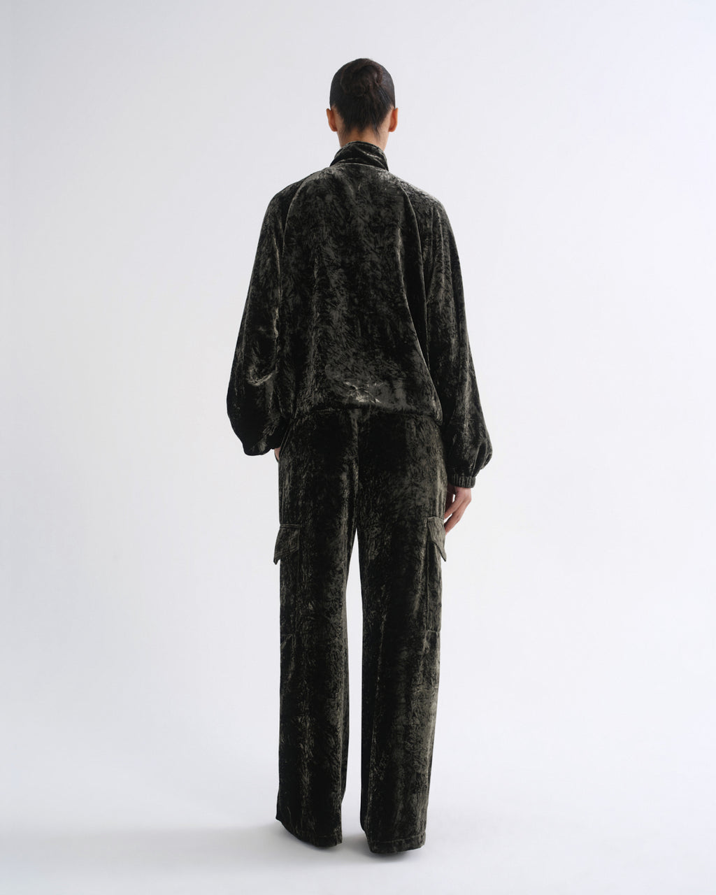 A model viewed from the back wearing a dark green crushed velvet two-piece set consisting of a long-sleeve high-neck jacket and matching wide-leg trousers, set against a plain light gray studio background.
