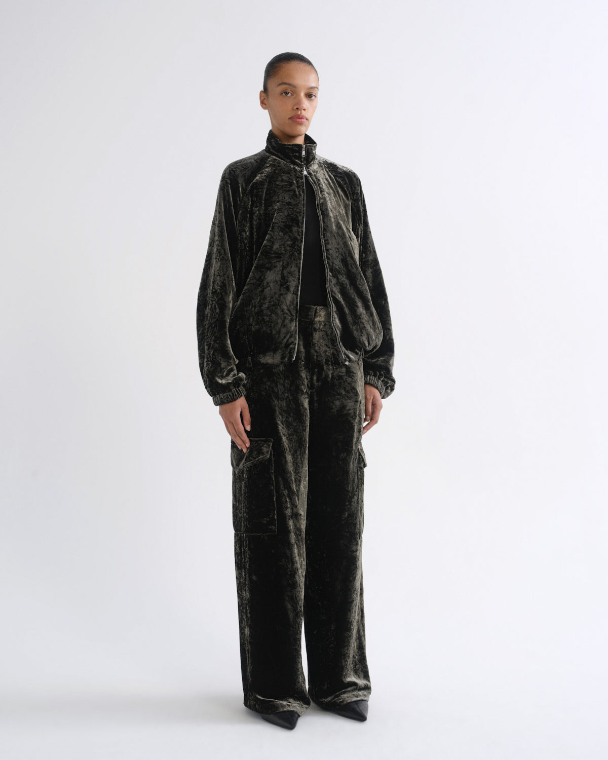 A model stands against a plain light gray studio background wearing a matching dark green velvet tracksuit with a zip-up jacket and wide-leg pants.