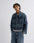 A man wearing a dark blue denim jacket and matching wide-leg denim jeans, shown from a side-back angle against a light gray studio background.