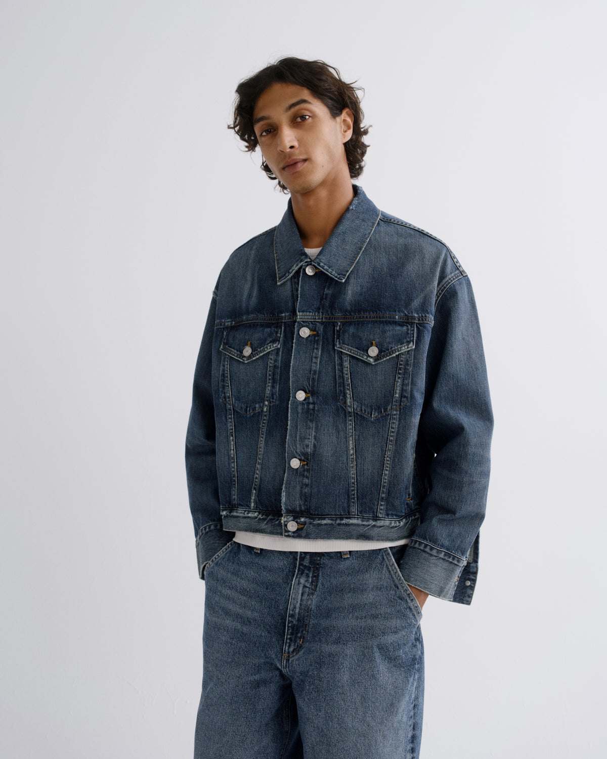 A man wearing a dark blue denim jacket and matching wide-leg denim jeans, shown from a side-back angle against a light gray studio background.