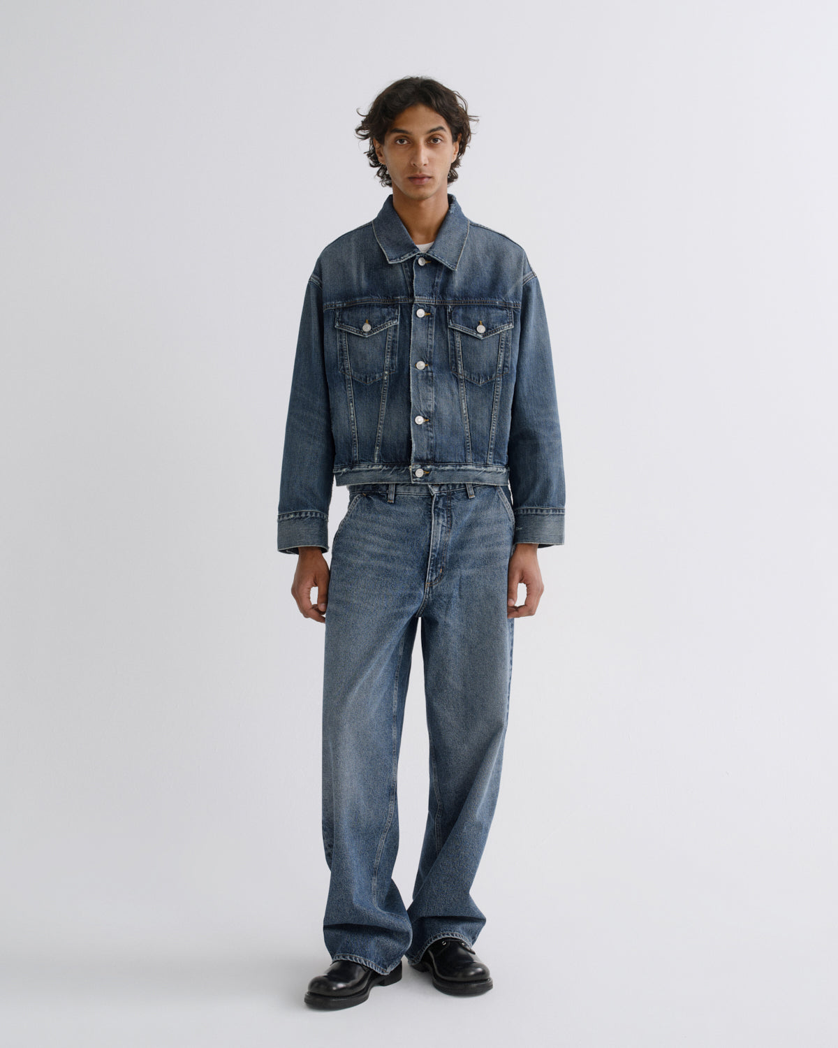 A model stands against a light gray studio background wearing a blue denim jacket and matching wide-leg denim jeans with black shoes.