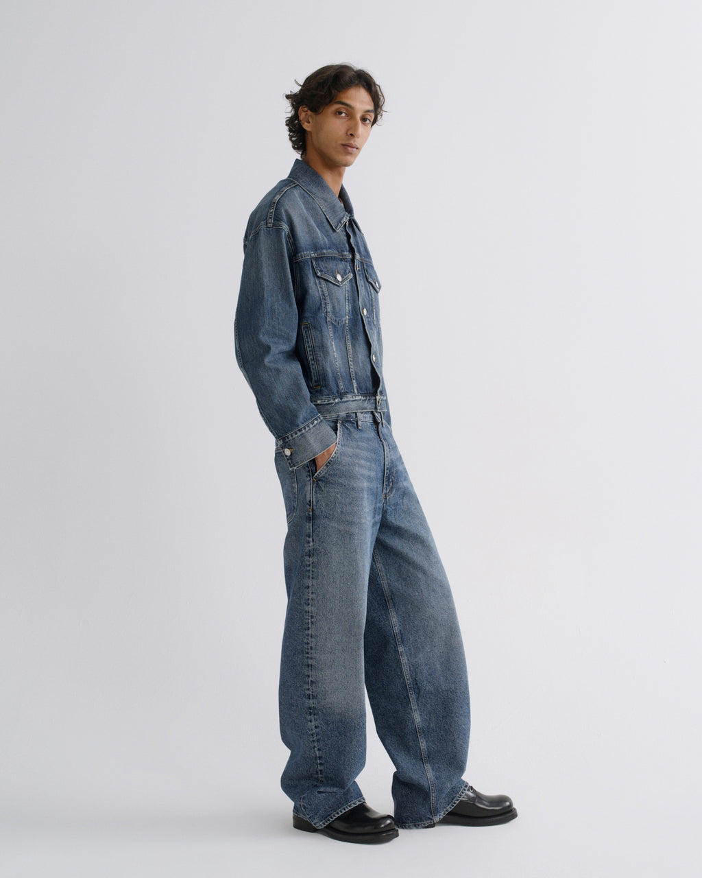 A model wearing a blue  denim jacket, styled with black leather shoes, standing in profile against a light gray studio background.