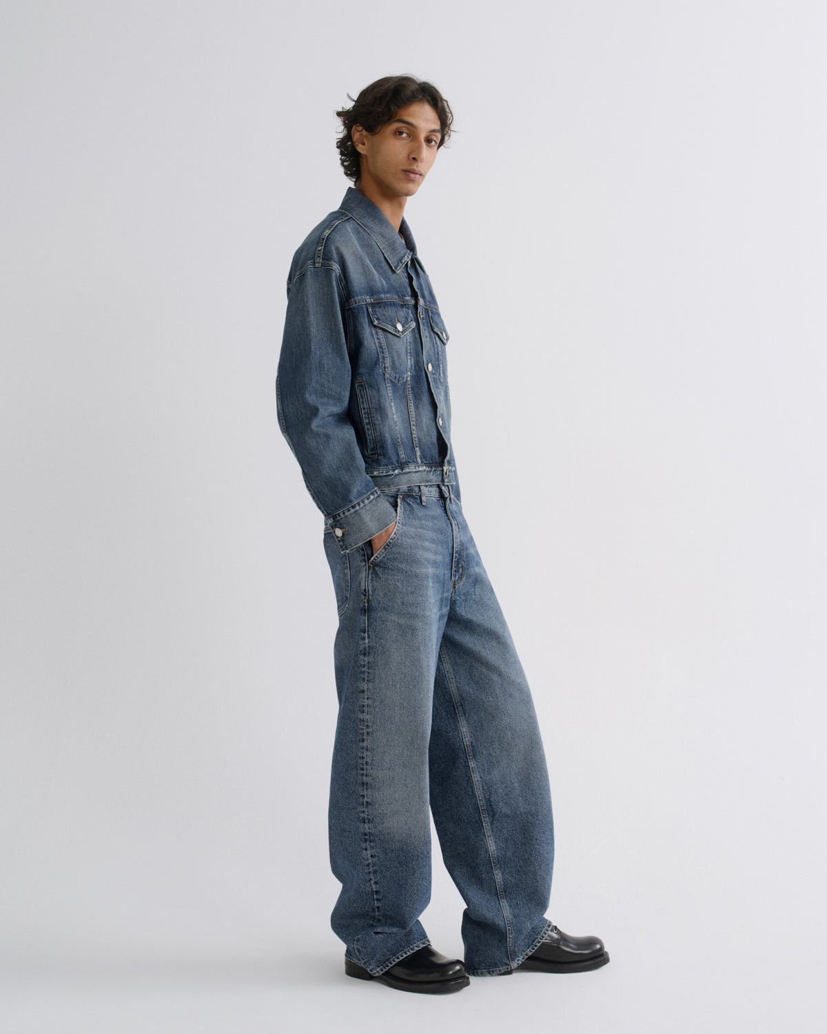 A model wearing a blue  denim jacket, styled with black leather shoes, standing in profile against a light gray studio background.