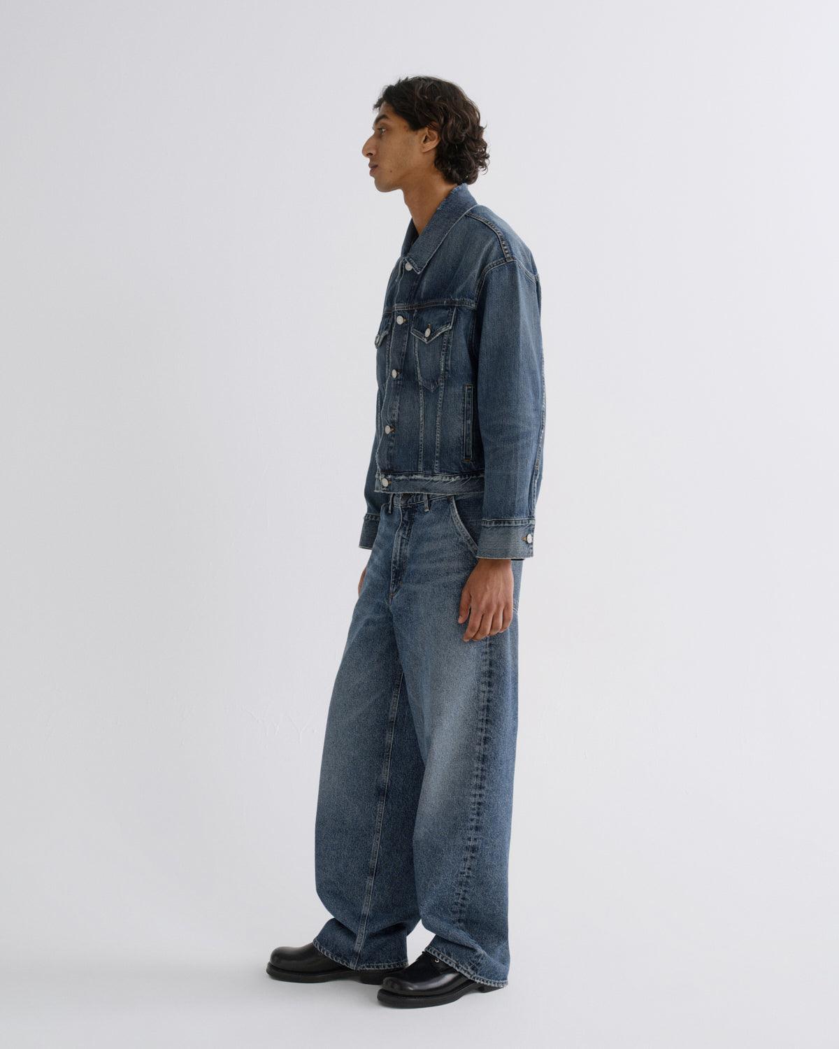 A man wearing a dark blue denim jacket and matching wide-leg denim jeans, standing in profile against a plain light gray studio background.