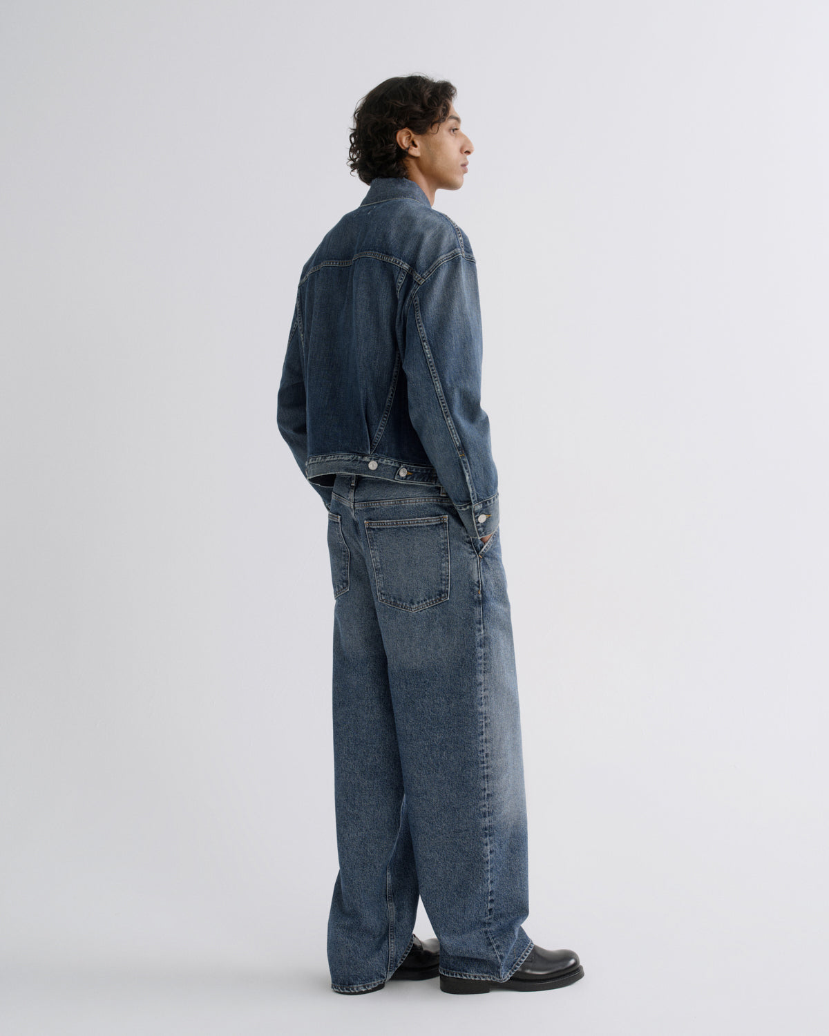 A man wearing a dark blue denim jacket and matching wide-leg denim jeans, shown from a side-back angle against a light gray studio background.