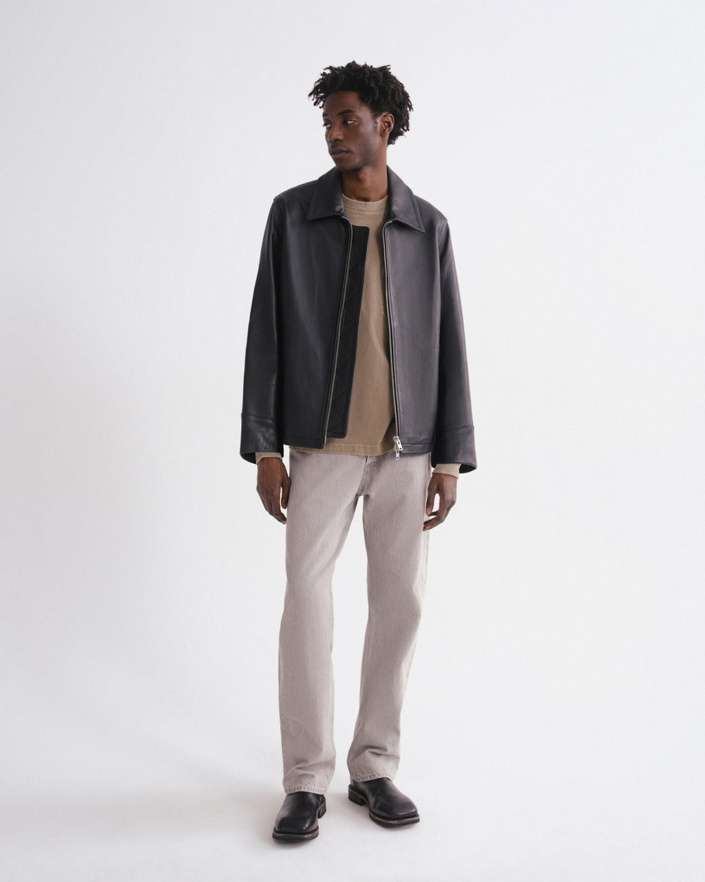 A man modeling a dark charcoal leather jacket over a beige crewneck sweater, paired with light beige trousers and black leather shoes, against a plain white studio background.