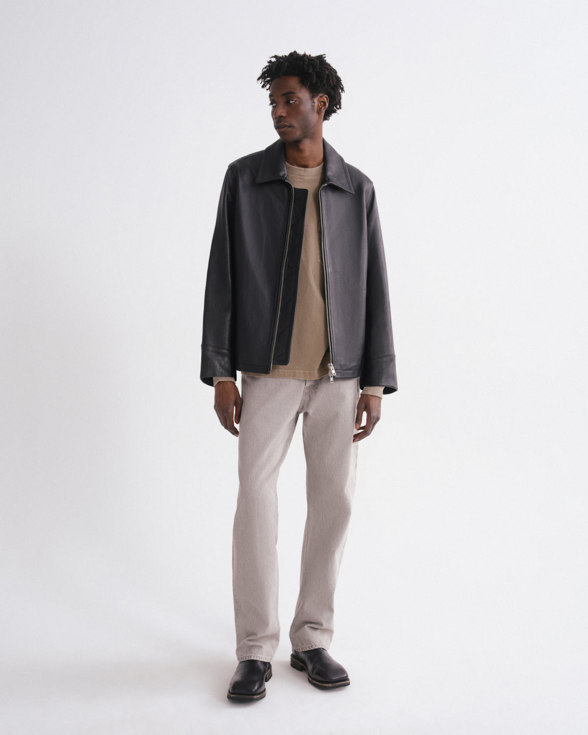 A man modeling a dark charcoal leather jacket over a beige crewneck sweater, paired with light beige trousers and black leather shoes, against a plain white studio background.
