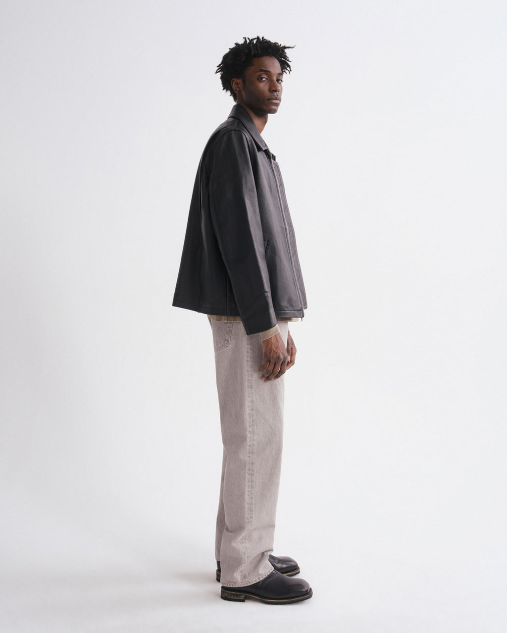 A man stands in profile wearing a dark charcoal gray oversized jacket, light beige wide-leg trousers, and black leather loafers against a plain white studio background.