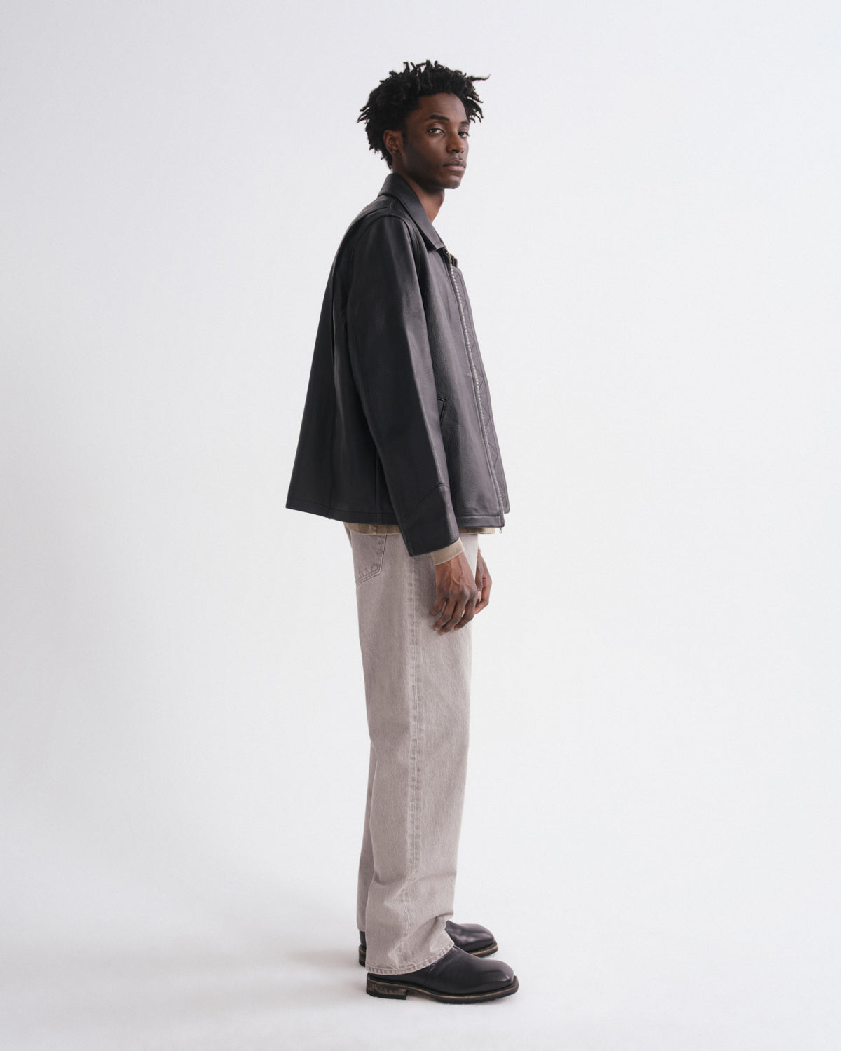 A man stands in profile wearing a dark charcoal gray oversized jacket, light beige wide-leg trousers, and black leather loafers against a plain white studio background.