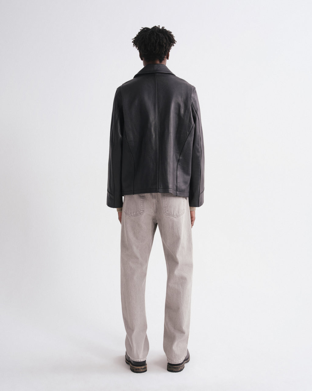 A man viewed from behind wearing a black leather jacket, light beige trousers, and dark shoes, standing against a plain white studio background.