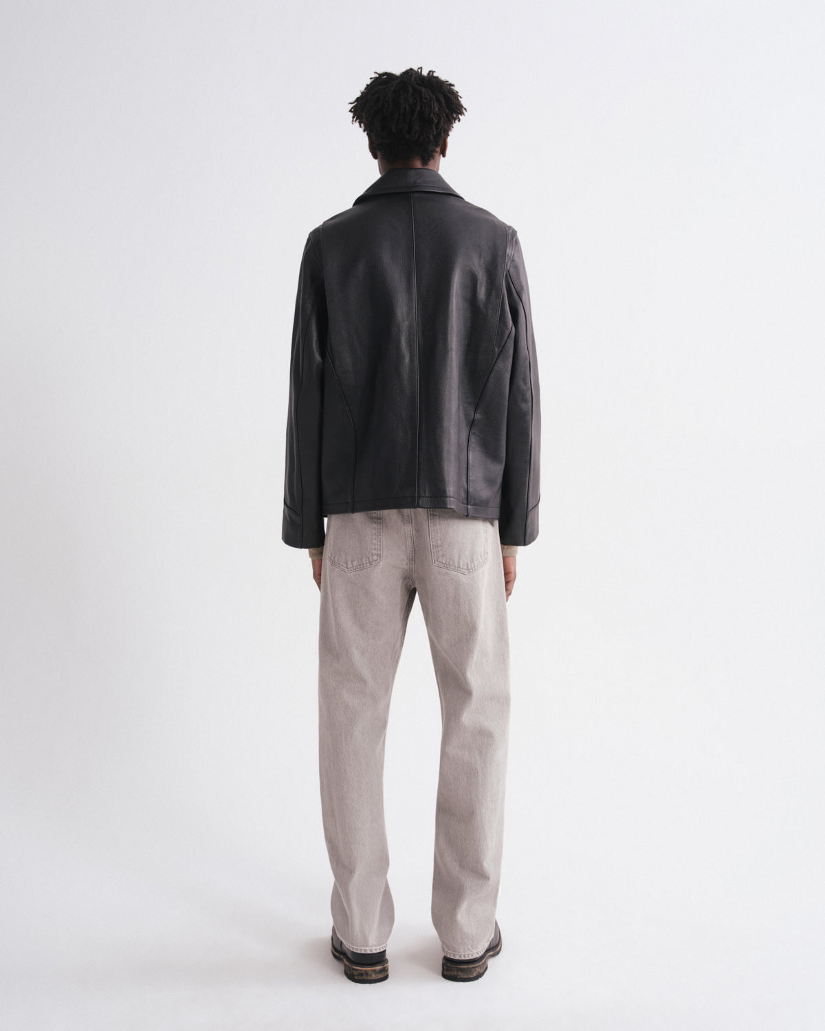 A man viewed from behind wearing a black leather jacket, light beige trousers, and dark shoes, standing against a plain white studio background.
