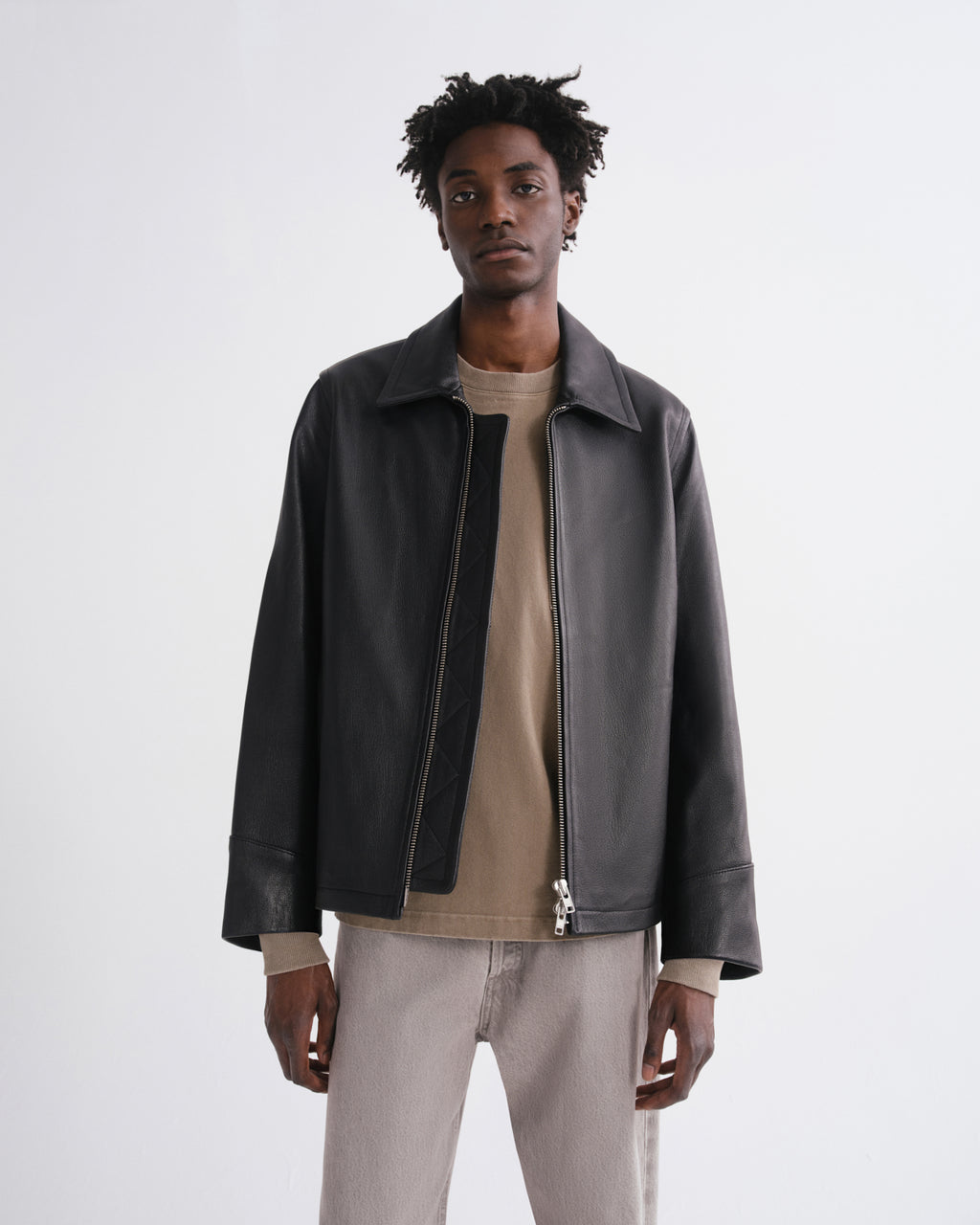 A man wearing a black leather jacket over a beige t-shirt and light gray trousers, standing against a plain light gray studio background.
