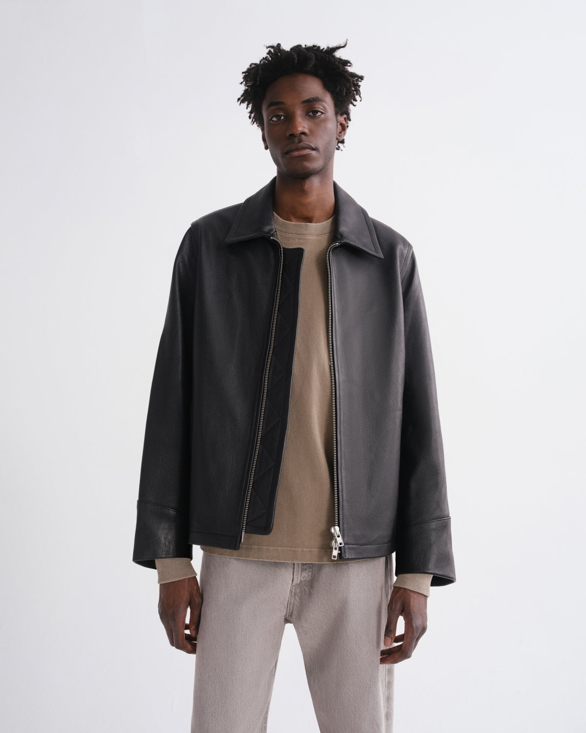 A man wearing a black leather jacket over a beige t-shirt and light gray trousers, standing against a plain light gray studio background.