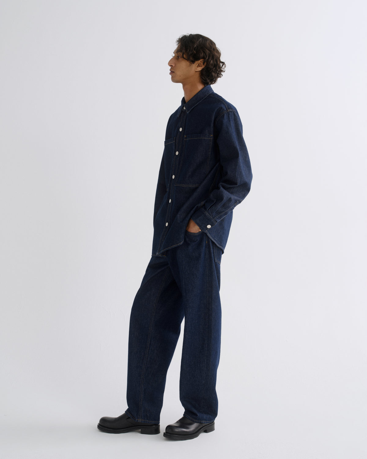 A man wearing a dark navy blue denim work jacket and matching wide-leg trousers, standing in profile against a light gray studio background.