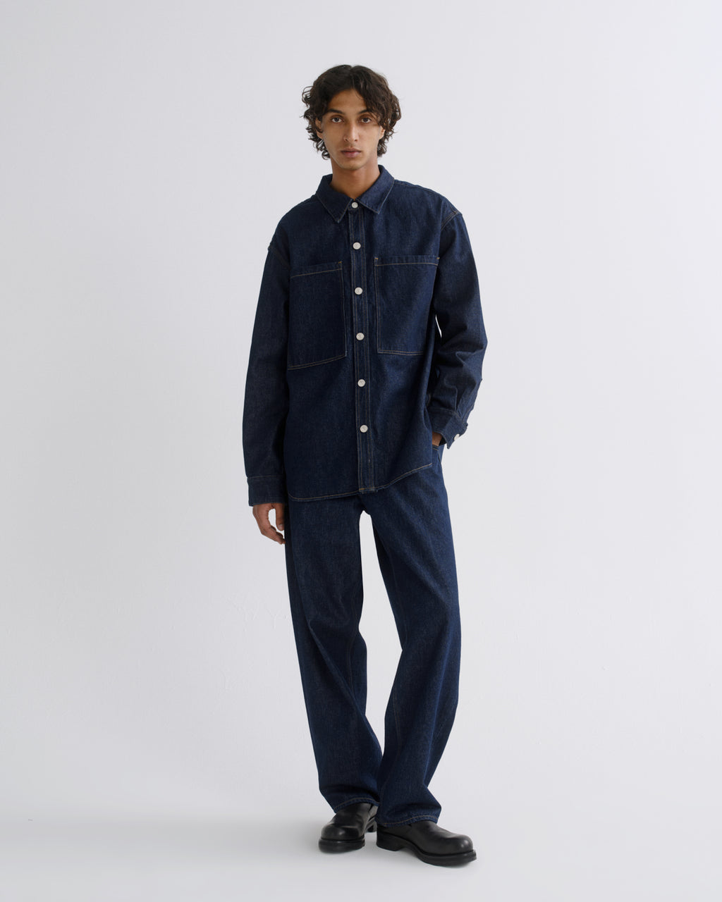 A model stands against a light gray studio background wearing a dark indigo denim work jacket and matching wide-leg denim pants, paired with black leather shoes.