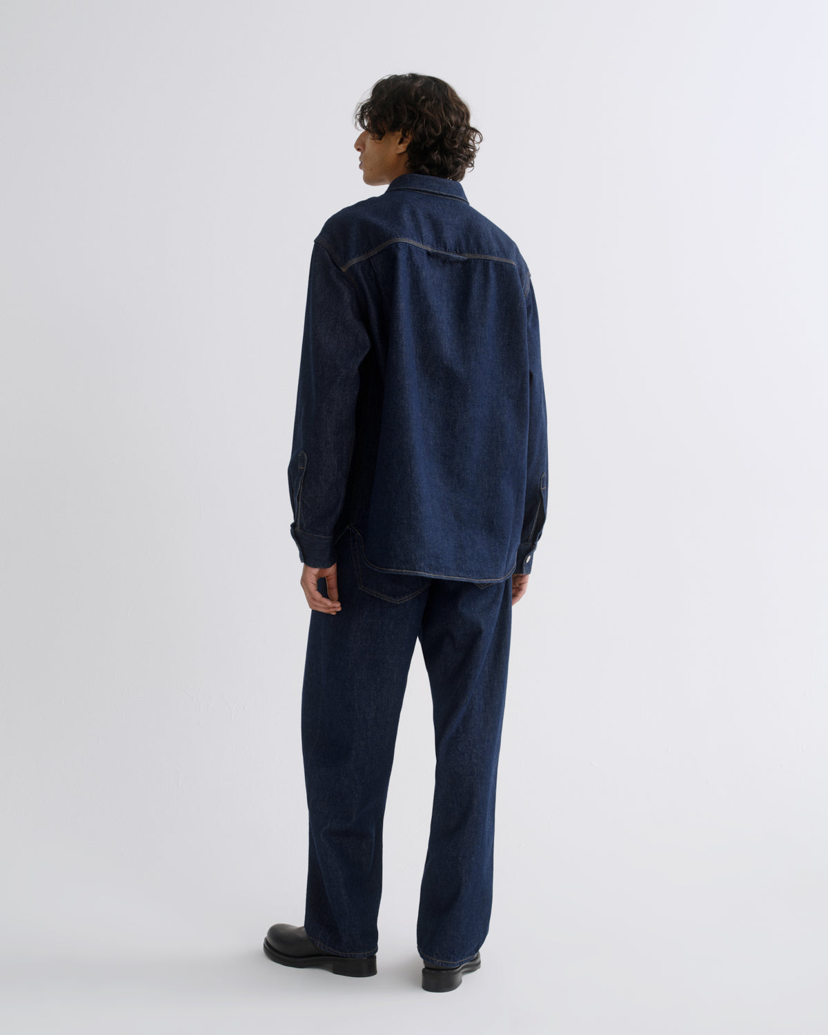 A model shown from the back wearing a dark indigo denim long-sleeve shirt and matching dark blue jeans against a plain light gray studio background.