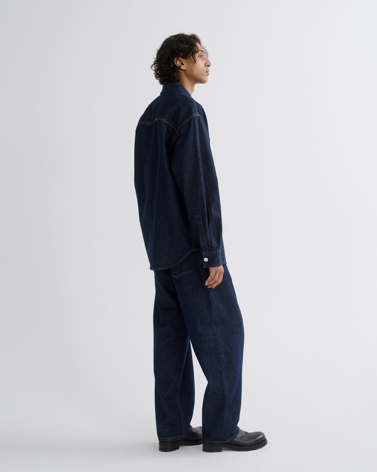 A man wearing a dark navy blue corduroy jacket and matching wide-leg jeans, shown from a side profile against a light gray studio background.