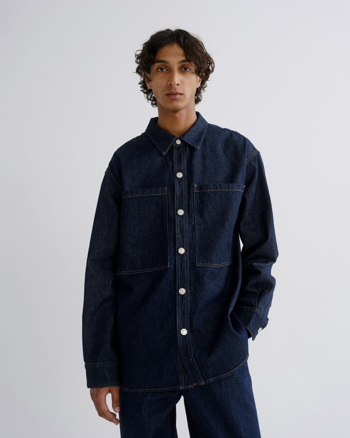 A man wearing a dark indigo denim button-up shirt jacket with front pockets, paired with matching dark denim pants, photographed against a light gray studio background.