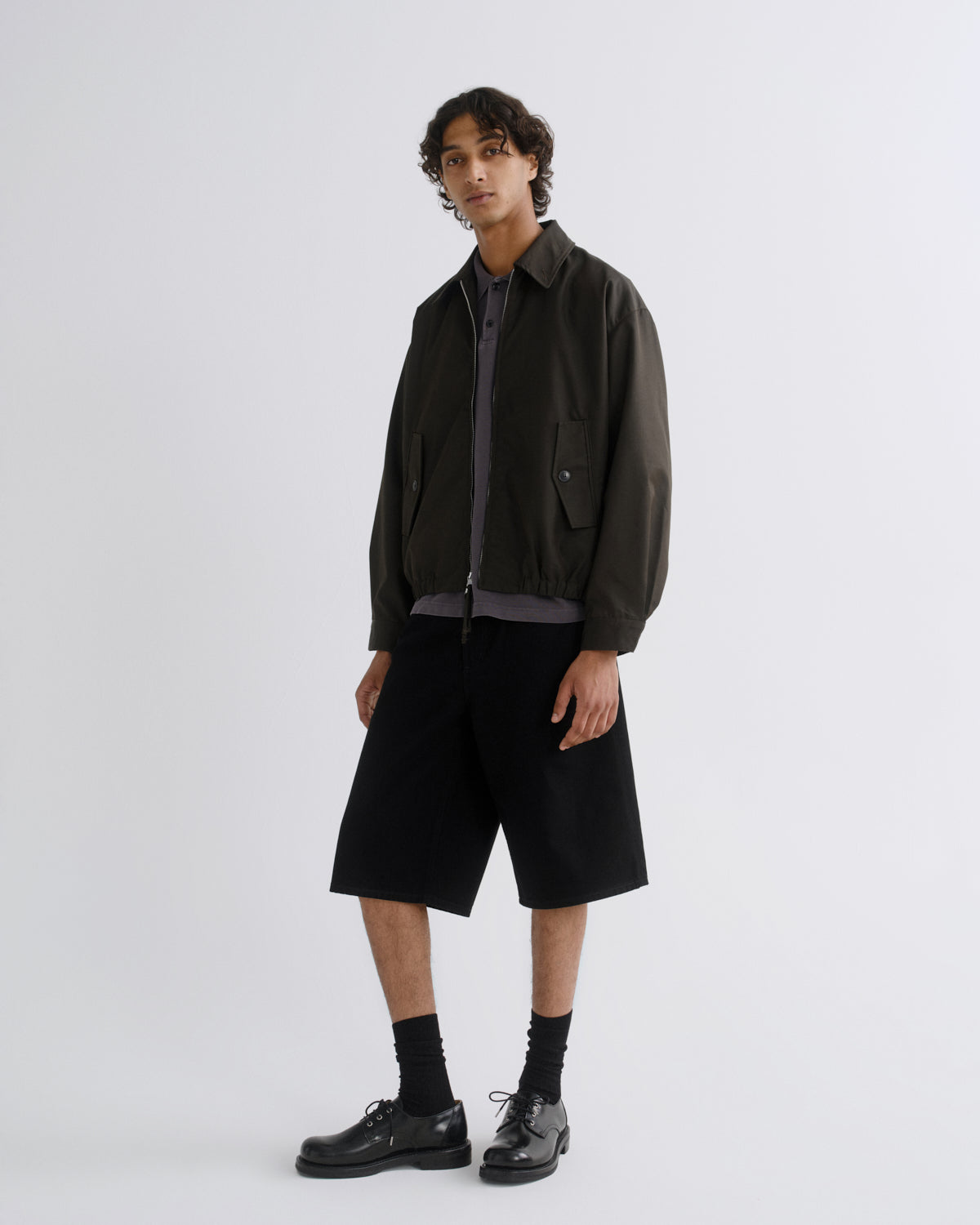 A model stands against a light gray studio background wearing a dark olive green utility jacket over a dark gray zip-up shirt, black wide-leg shorts, black socks, and black leather shoes.