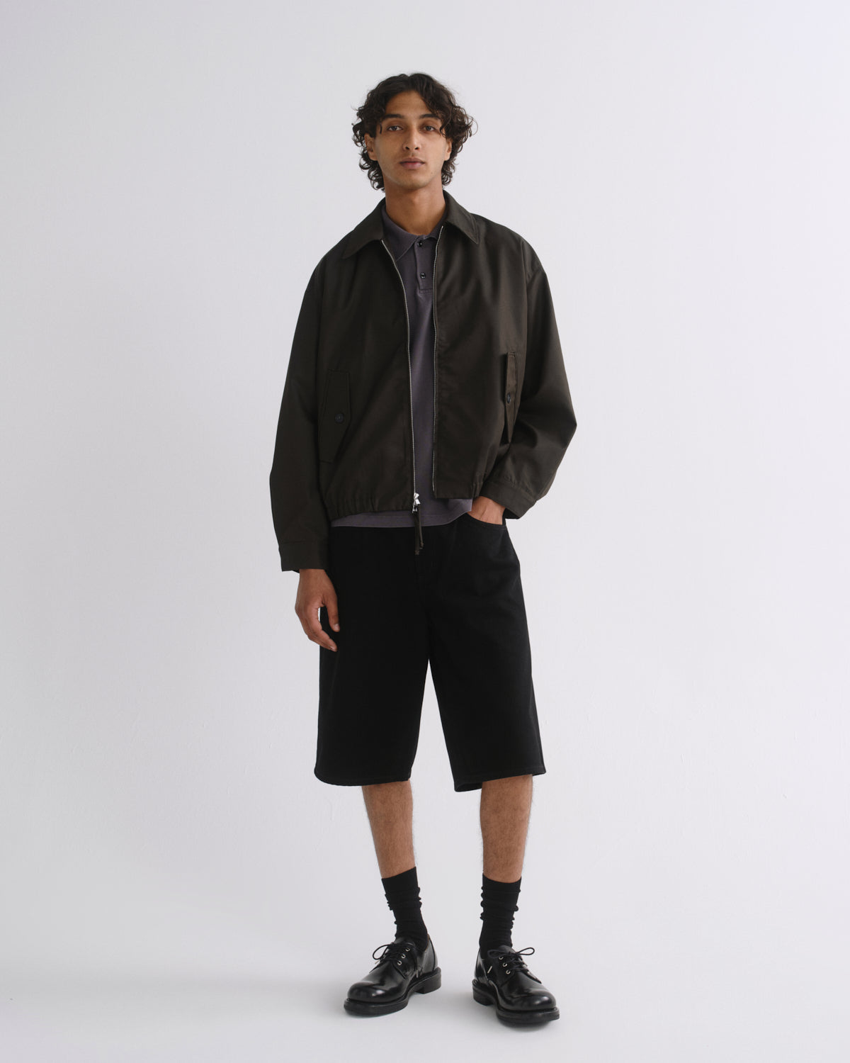 A model stands against a light gray studio background wearing a dark olive-brown jacket over a dark gray zip-up shirt, black shorts, black socks, and black leather shoes.
